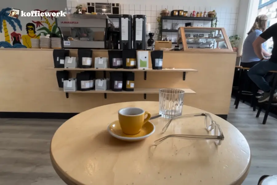 At Tigershark in The Hague: yellow espresso cup on a round table with a water glass and glasses case, coffee counter and bags in the background.
