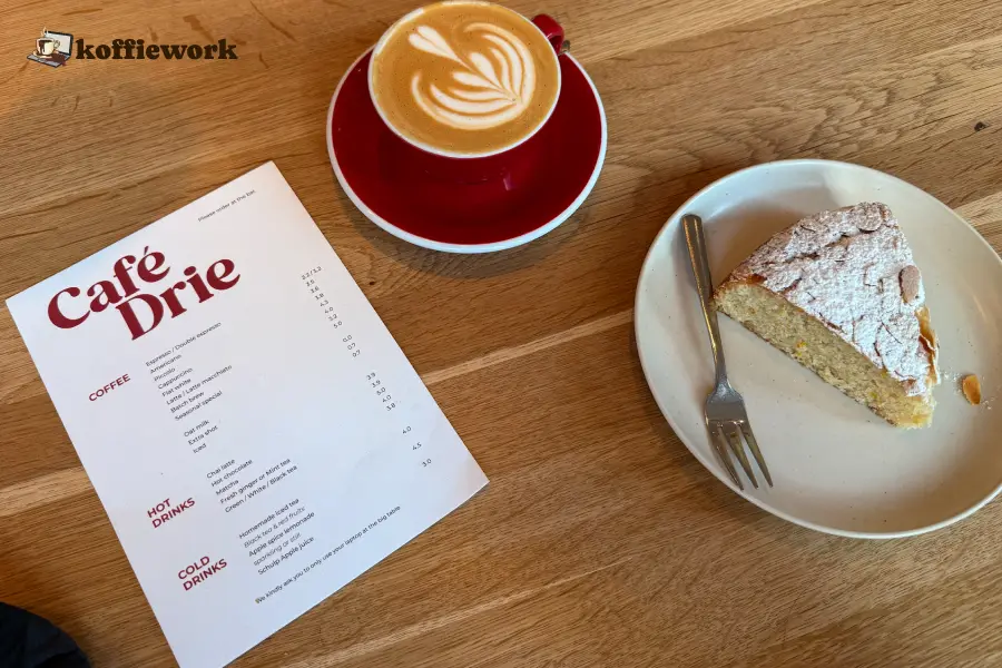 Cafe Drie in The Hague: latte with leaf latte art next to the Cafe Drie menu and a slice of cake on a plate.